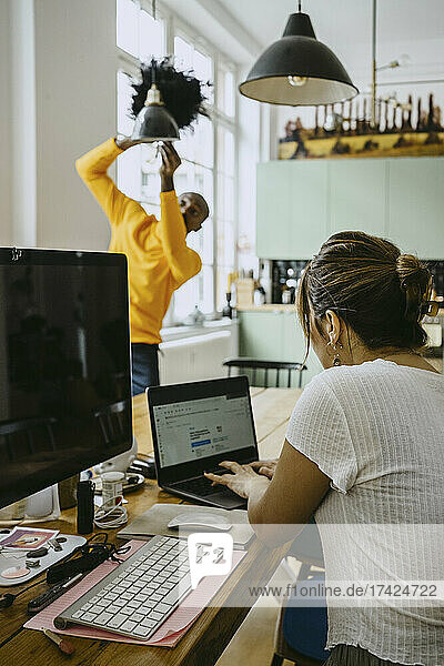 Eine Freiberuflerin benutzt einen Laptop,  während ein Mann eine Pendelleuchte zu Hause reinigt
