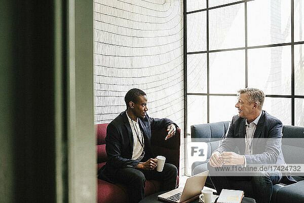 Männlicher Unternehmer im Gespräch mit einem reifen Geschäftsmann  der auf einem Sofa im Kongresszentrum sitzt