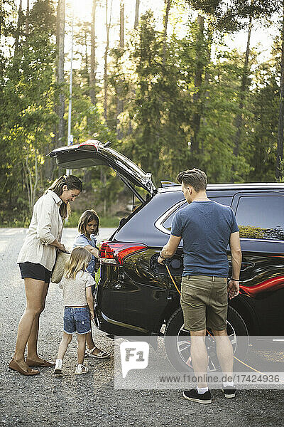 Porträt einer Mutter  eines Vaters und zweier Töchter  die neben einem Auto an einer Ladestation für Elektrofahrzeuge stehen