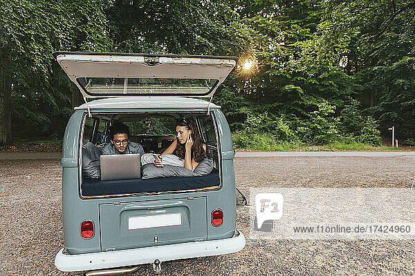 Junger Mann mit Laptop  während seine Freundin mit Handy im Campingwagen liegt