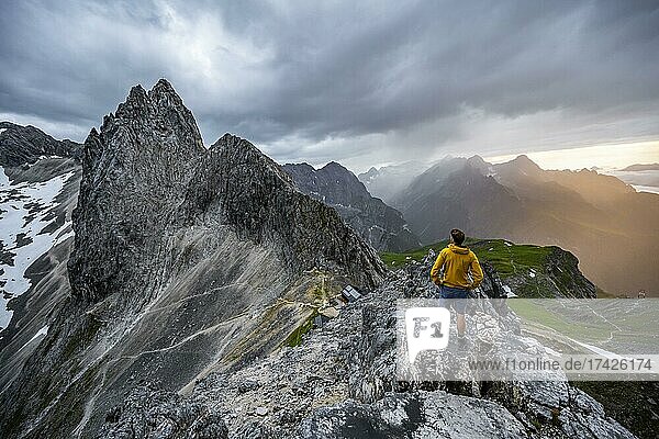 Hikers at the summit of the Westliche Törlspitze  in the background cloudy mountains at sunset with dramatic light  craggy rocky summit of the Partenkirchener Dreitorspitze  in the background Reintal and Zugspitzplatt  Wetterstein Mountains  Garmisch Partenkirchen  Bavaria  Germany  Europe
