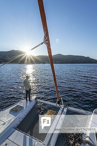 Junger Mann steht auf einem Katamaran und blickt in den Sonnenuntergang  Seile auf einem Segel-Katamaran  Sonnenstern  Segeltörn  Tilos  Dodekanes  Griechenland  Europa