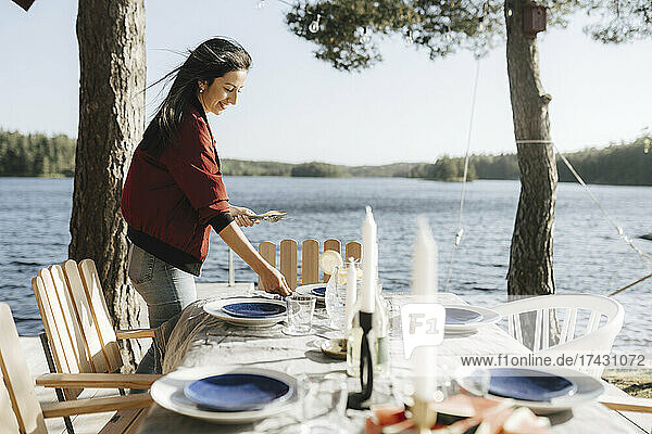Smiling young woman setting table by lake on sunny day