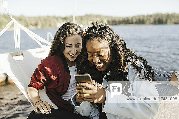 Lächelnde Freundinnen benutzen ihr Smartphone  während sie in der Hängematte sitzen