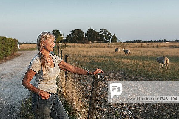 Lächelnde ältere Bäuerin  die am Zaun steht und auf ein Feld schaut