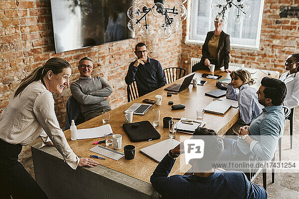 Reife Geschäftsfrau  die sich auf einen Konferenztisch stützt  während sie mit Kollegen im Sitzungssaal einen Plan bespricht
