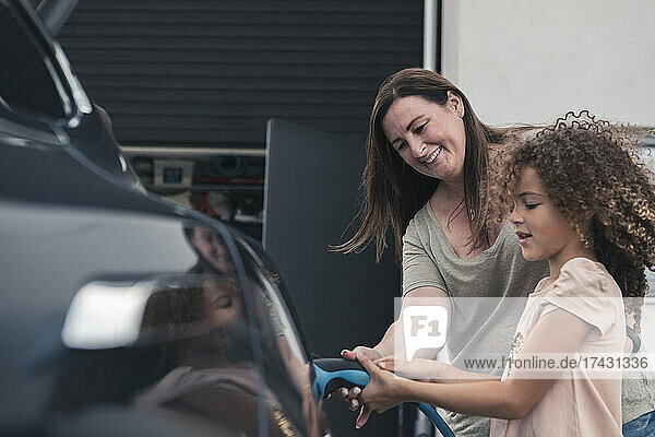 Lächelnde Mutter  die ihrer Tochter beim Aufladen eines Elektroautos im Vorgarten hilft