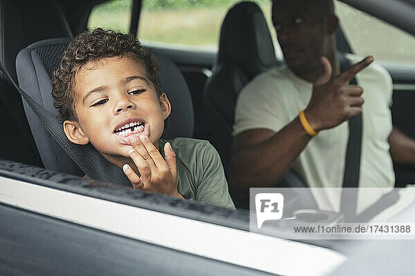 Playful boy showing broken teeth while sitting by father in electric car