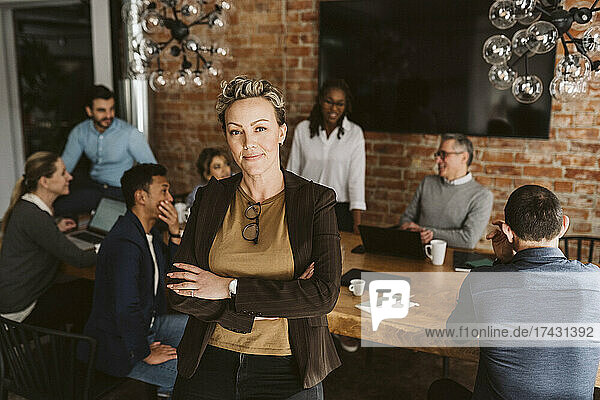 Portrait of smiling businesswoman standing with arms crossed while colleagues discussing in background