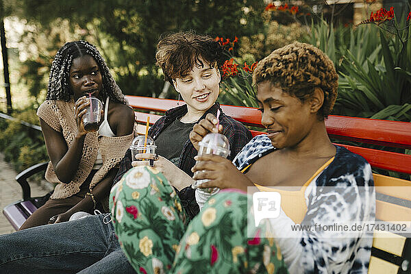Junge Frau und Teenager schauen sich einen nicht-binären Freund beim Trinken im Park an