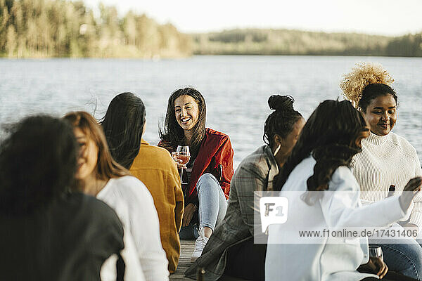 Smiling multiracial female friends sitting on jetty against lake