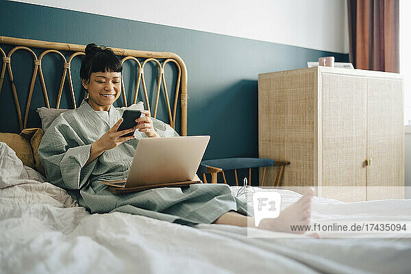 Lächelnde Frau mit Laptop und Smartphone auf dem Bett zu Hause
