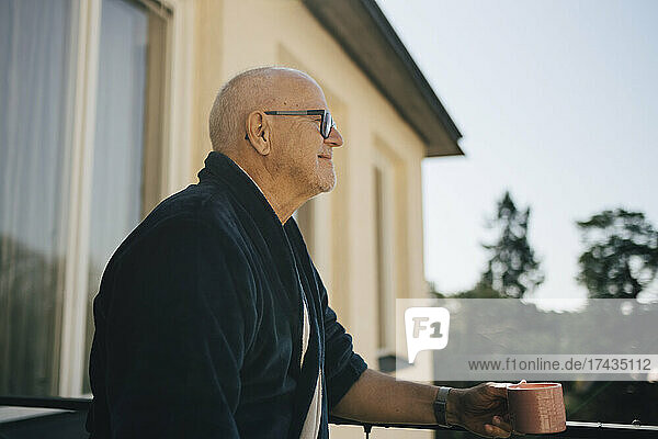 Lächelnder älterer Mann schaut weg  während er eine Kaffeetasse auf dem Balkon hält