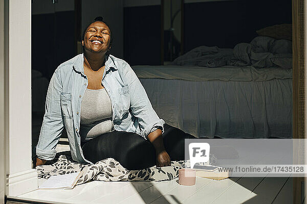 Lächelnde Frau sitzt mit geschlossenen Augen im Schlafzimmer an einem sonnigen Tag