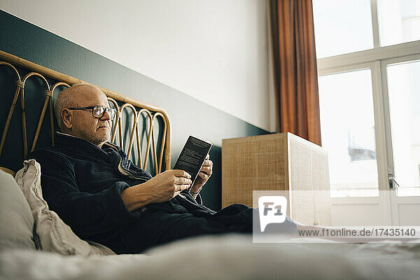 Älterer Mann liest ein Buch  während er auf dem Bett im Schlafzimmer sitzt