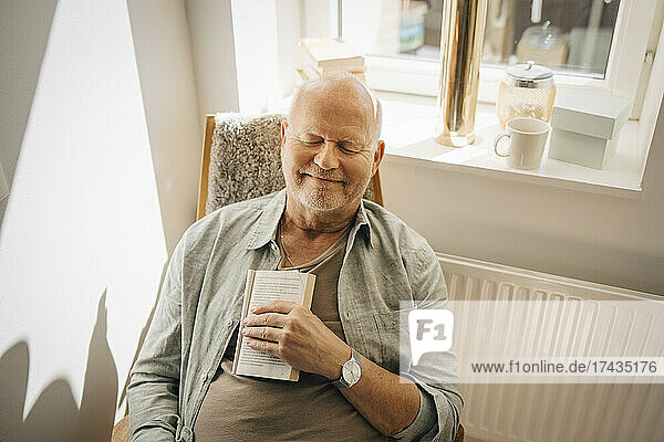 Lächelnder älterer Mann mit Buch  der sich auf einem Stuhl zu Hause entspannt