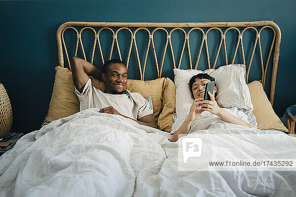 Lächelnde Freundin teilt ihr Smartphone mit ihrem Freund  während sie auf dem Bett liegt