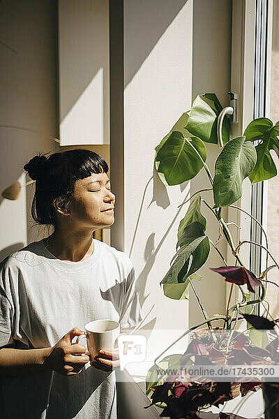 Lächelnde Frau hält eine Kaffeetasse  während sie mit geschlossenen Augen im Sonnenlicht zu Hause steht