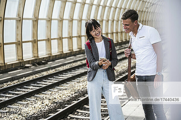 Happy male and female friends sharing mobile phone at train station platform