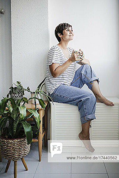 Mid adult woman with mug sitting near plants at home