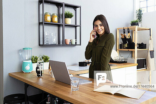 Businesswoman talking on smart phone while standing in kitchen at home