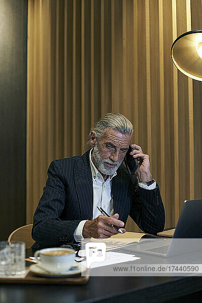 Männlicher Berufstätiger  der am Schreibtisch im Hotel sitzt und auf dem Smartphone spricht
