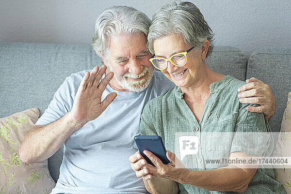 Älterer Mann winkt während eines Videoanrufs über sein Smartphone  während er zu Hause neben einer Frau sitzt
