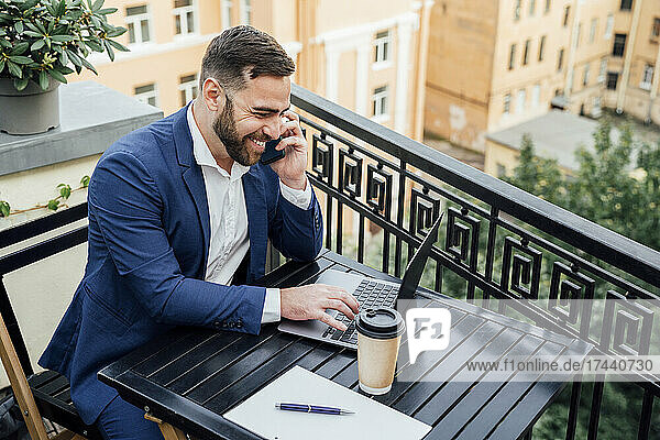 Smiling businessman using laptop while talking on smart phone in balcony