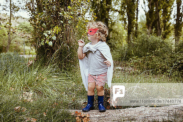 Boy with blond hair wearing cape while standing on log in forest