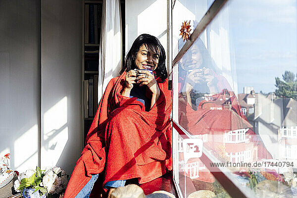 Lächelnde Frau mit Schal beim Kaffeetrinken am Fenster zu Hause