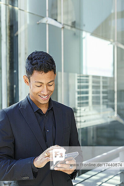 Smiling young businessman surfing net through mobile phone while standing in front of office building