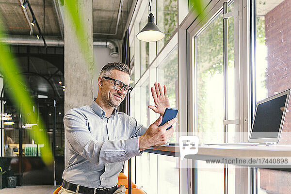 Männlicher Berufstätiger winkt während eines Videoanrufs über das Mobiltelefon im Büro mit der Hand
