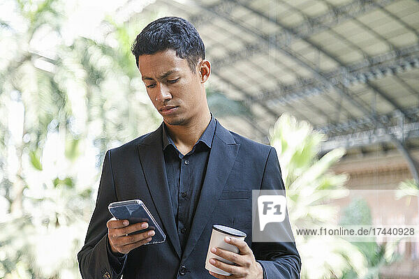 Male professional holding disposable coffee cup while using smart phone