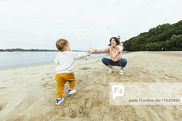 Male toddler walking towards mother crouching on sand at beach