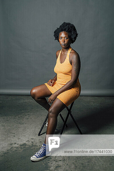 Afro young woman wearing orange dress sitting on chair in studio