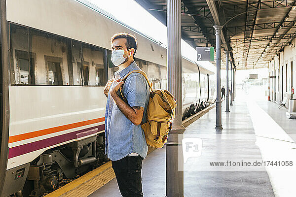 Young man with backpack standing on railroad station platform during pandemic