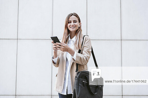 Smiling businesswoman with mobile phone and shoulder bag in front of white wall