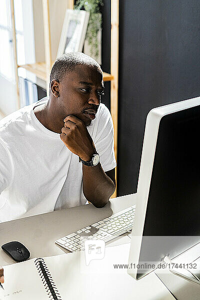Male freelancer sitting with hand on chin looking at desktop computer in studio