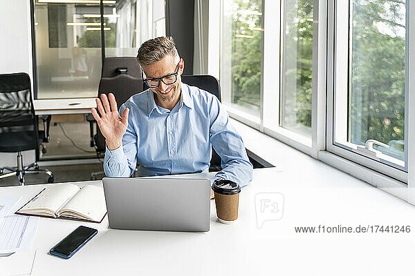 Smiling businessman waving hand during video call through laptop at desk