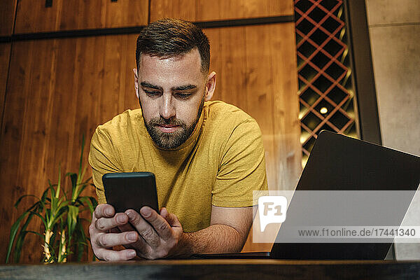 Male freelancer using mobile phone while leaning on desk at home
