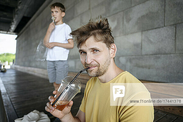 Man drinking iced coffee by son on bench