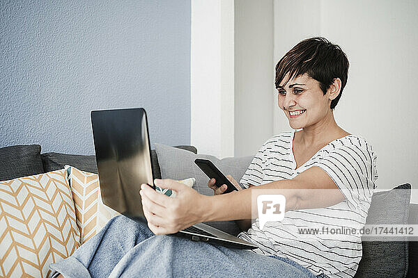 Lächelnde Freiberuflerin mit Smartphone und Laptop während eines Videoanrufs zu Hause