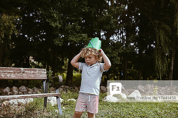 Cute blond boy wearing bucket on head standing by bench at backyard