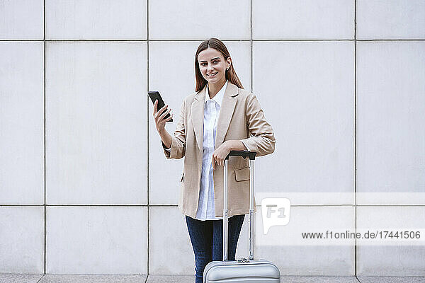 Smiling businesswoman with mobile phone and luggage in front of white wall