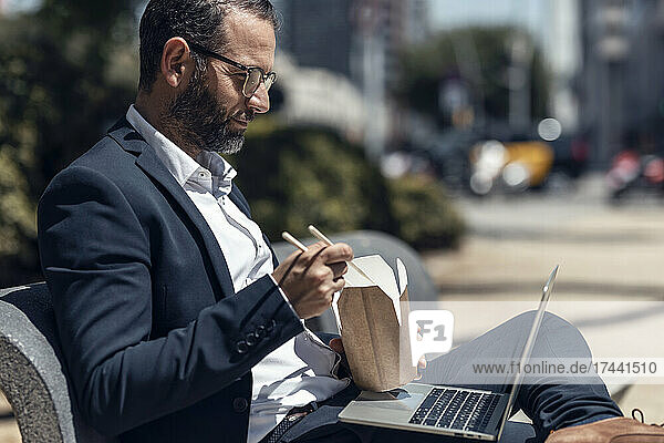 Businessman with laptop eating food while sitting on bench