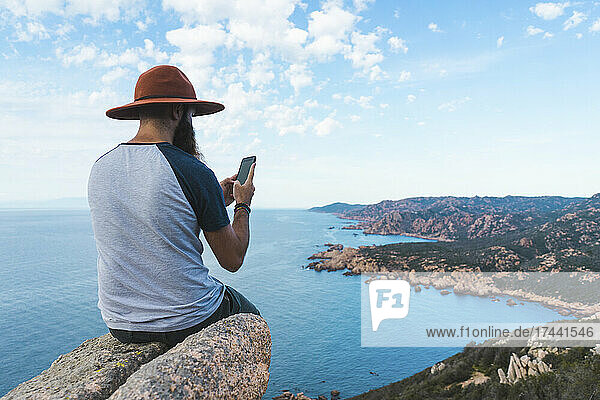 Junger Mann benutzt Mobiltelefon  während er auf einem Felsen sitzt