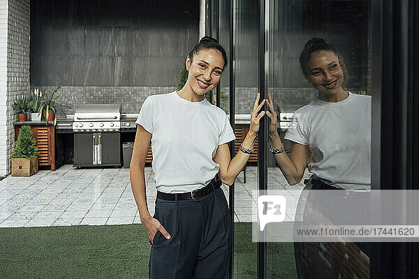 Smiling female professional with hand in pocket standing by glass wall