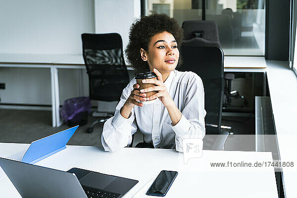 Businesswoman holding disposable coffee cup while sitting with laptop in office