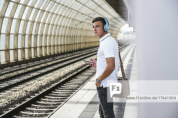 Young man with mobile phone and shoulder bag standing at train station