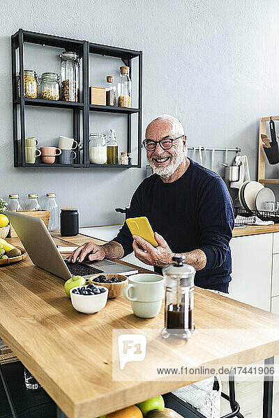 Lächelnder älterer Mann mit Mobiltelefon und Laptop sitzt am Küchentisch